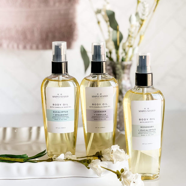 Three bottles of Simply Stated Bath & Body Aromatherapy Body Oil in Eucalyptus + Spearmint, Lavender + Vanilla, and Rosemary + Eucalyptus, arranged on a white tray with elegant floral details and a neutral, spa-inspired backdrop.