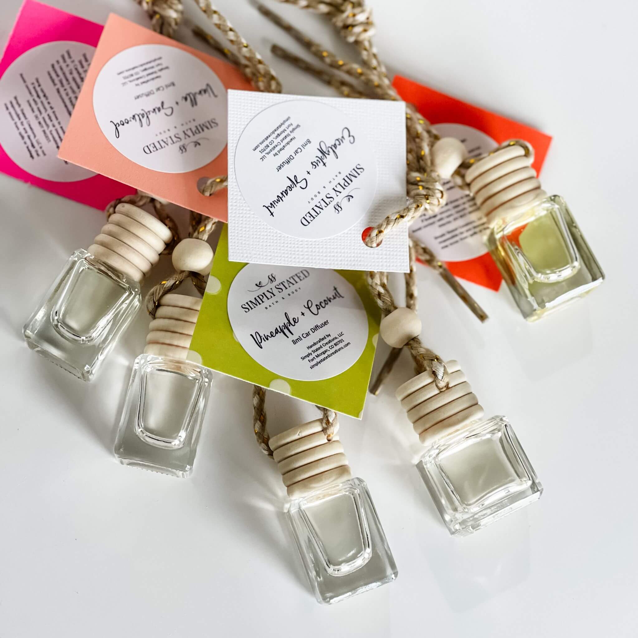 A collection of car diffusers with glass jars and wooden lids, attached to strings for hanging, displayed on a white surface with colorful labels indicating different scents.