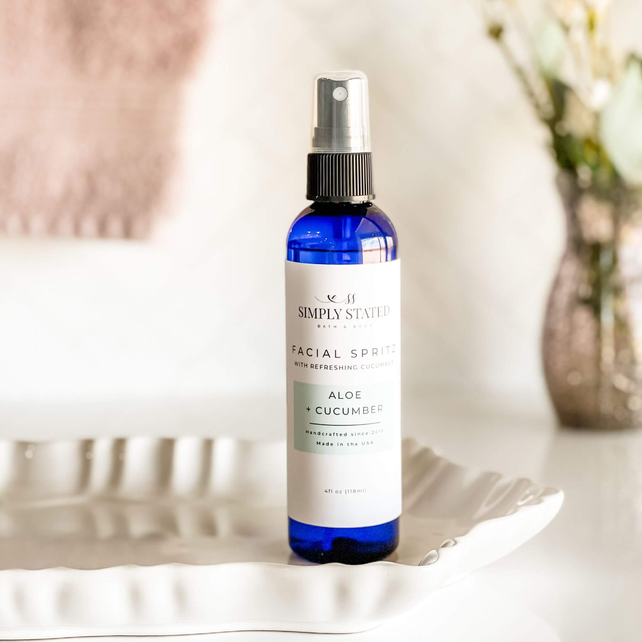 Simply Stated Facial Spritz in Aloe + Cucumber, refreshing facial mist in blue bottle placed on a white decorative dish with soft background decor.