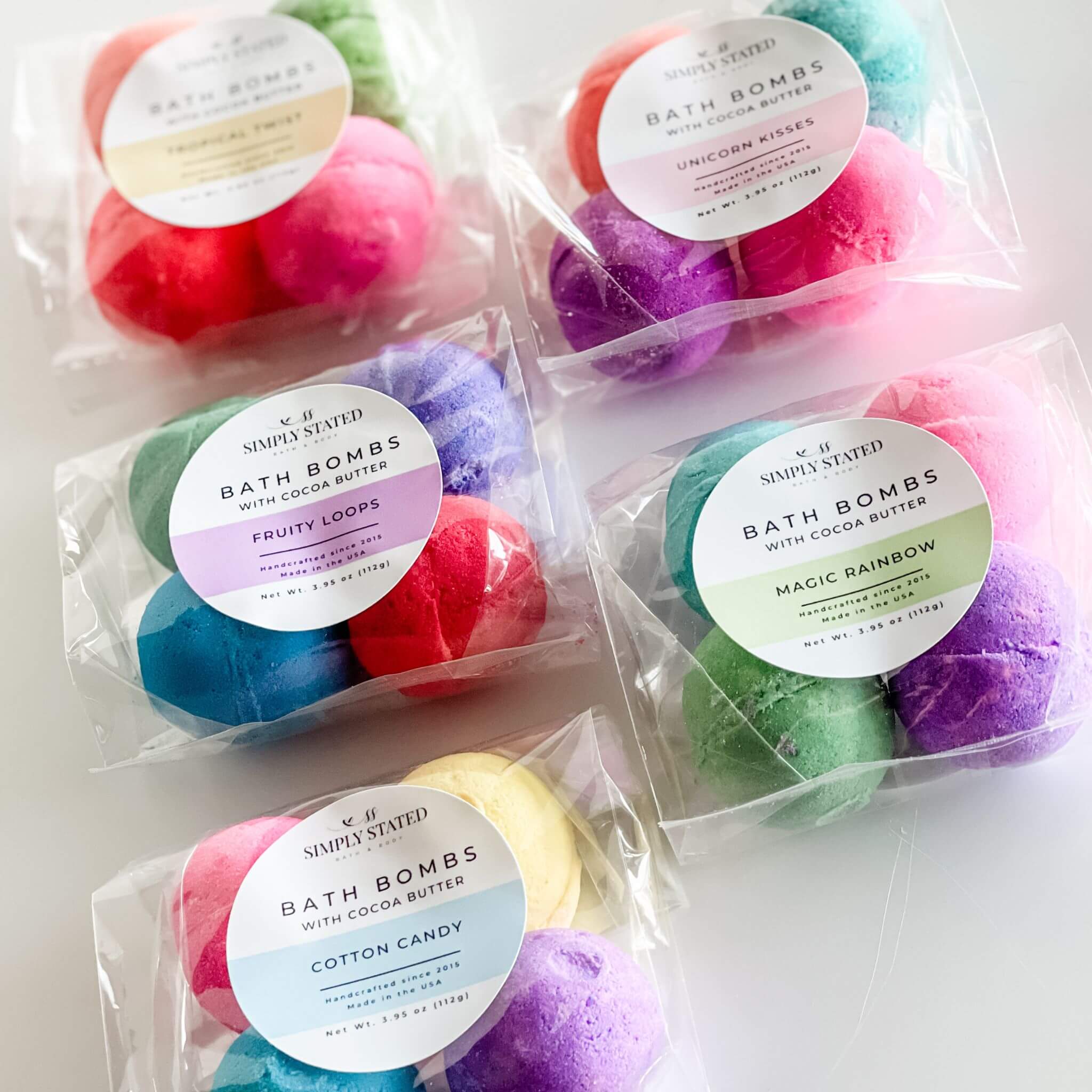 Group photo of Simply Stated Kids Line Bath Bomb Packs in various scents: Cotton Candy, Fruity Loops, Magic Rainbow, Tropical Twist, and Unicorn Kisses, arranged in vibrant, colorful packaging.