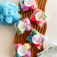 Packaged bath bombs on a wooden tray with a blue loofah and green towel.