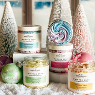 Collection of whipped soap jars with colorful labels and decorative Christmas trees in the background.