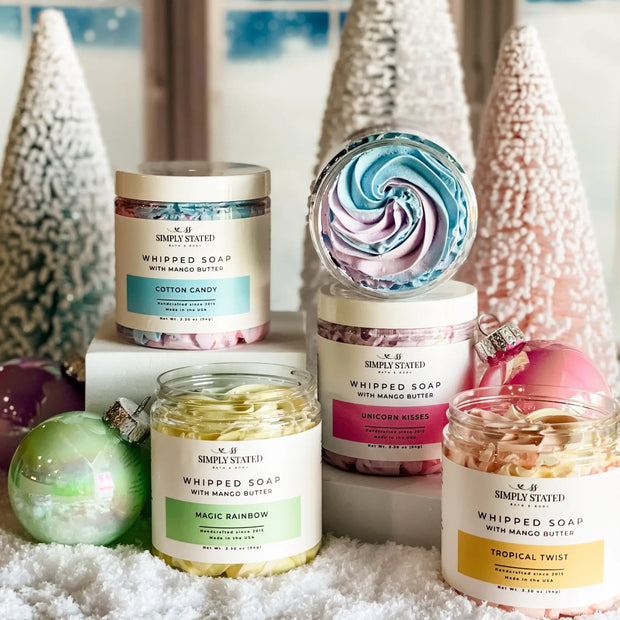 Collection of whipped soap jars with colorful labels and decorative Christmas trees in the background.