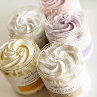 Open jars of Signature Line Whipped Soaps showcasing creamy whipped textures in vibrant colors, including Coconut Milk + Mango, Sweet Berry + Vanilla, Warm Vanilla + Sugar, Oatmeal Milk + Honey, and Sea Salt + Orchid.