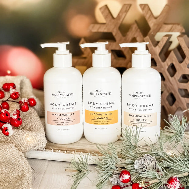 Three bottles of 'Simply Stated' body cream with Christmas decorations in the background