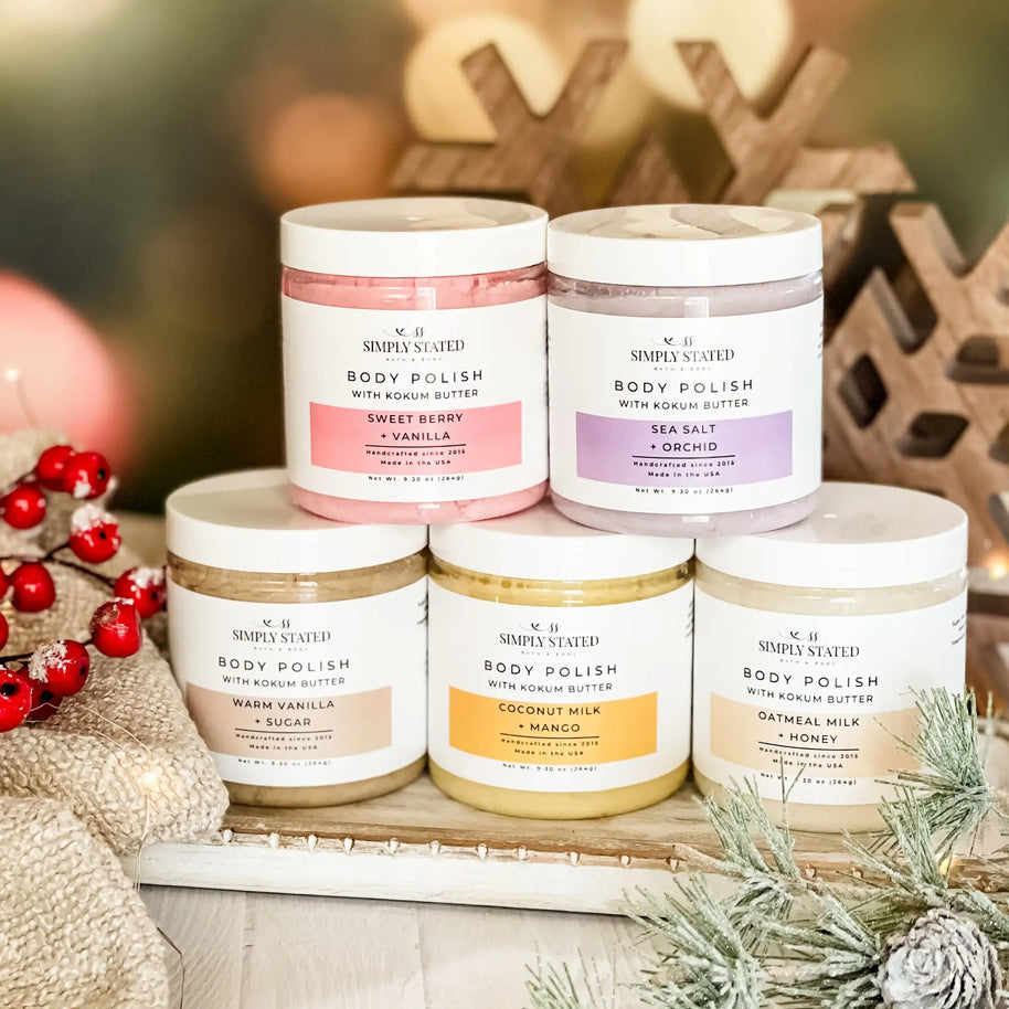 Five jars of body polish with different scents on a decorative surface with Christmas decorations.