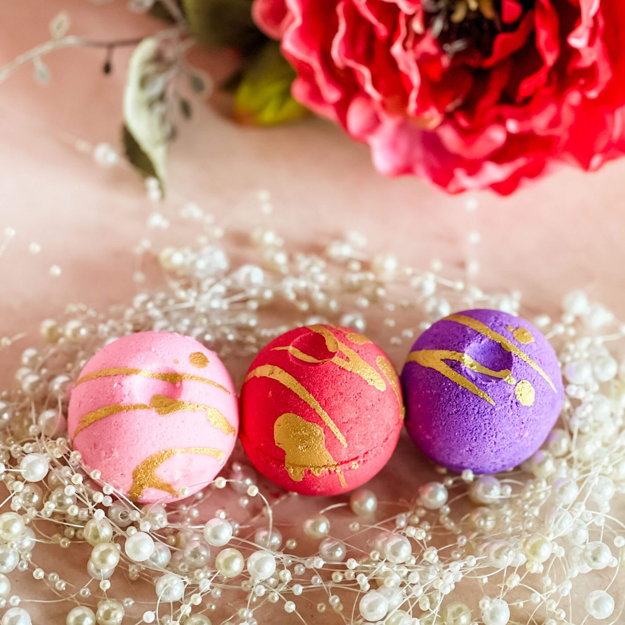 A trio of round bath bombs in pink, red, and purple with gold drizzle accents, displayed on

a soft pink background with pearl decor, representing the Wild Berry Kiss, Raspberry Vanilla Crush, and Strawberry Bubbly fragrances from the Sweet Temptations Collection.